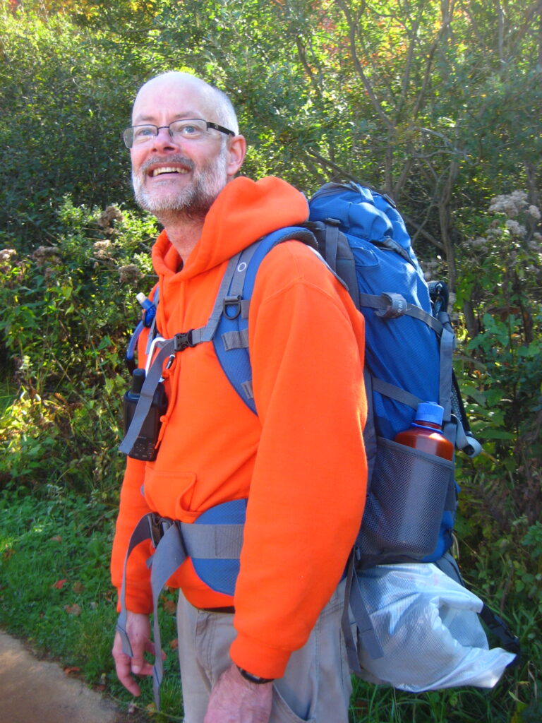 David Buskirk wearing a bright orange hoodie with a blue backpack on his back looking up in the sky. In the background are trees.