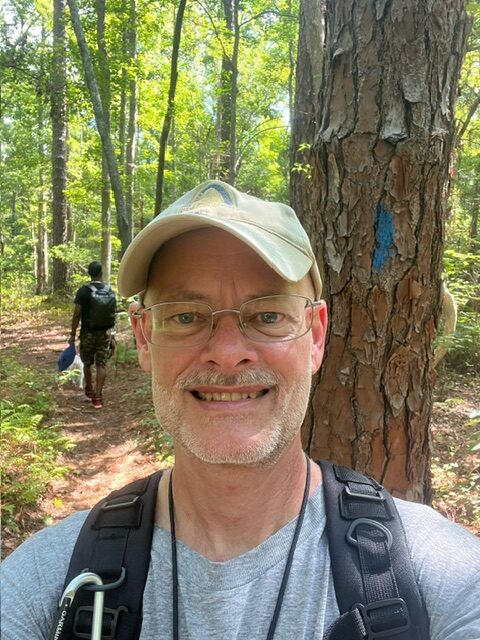 A man taking a selfie in the woods with a pine tree right behind him.